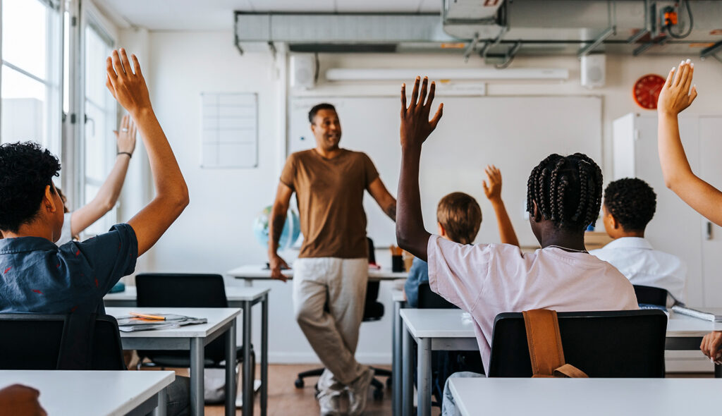 A teacher stands at the front of a class looking out on a classroom of students with their hands raised.