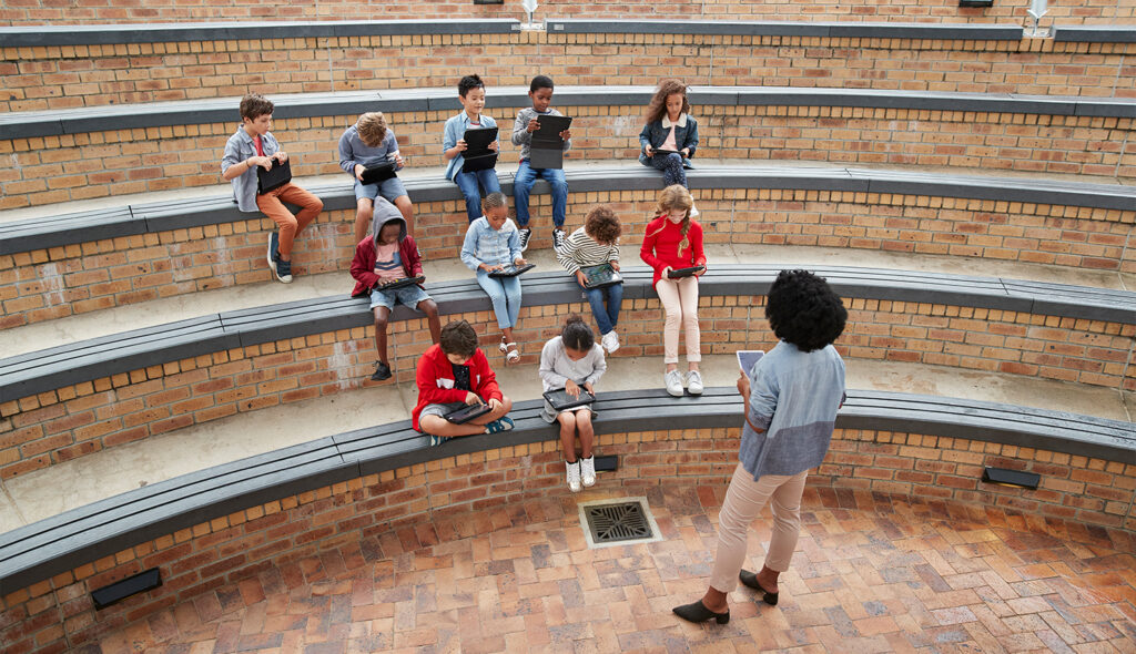 A teacher holds a tablet and speaks to her elementary school students in an outdoor classroom.