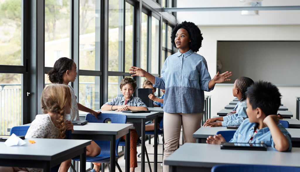 A teacher speaks to her class as she walks between rows of students seated in their desks.