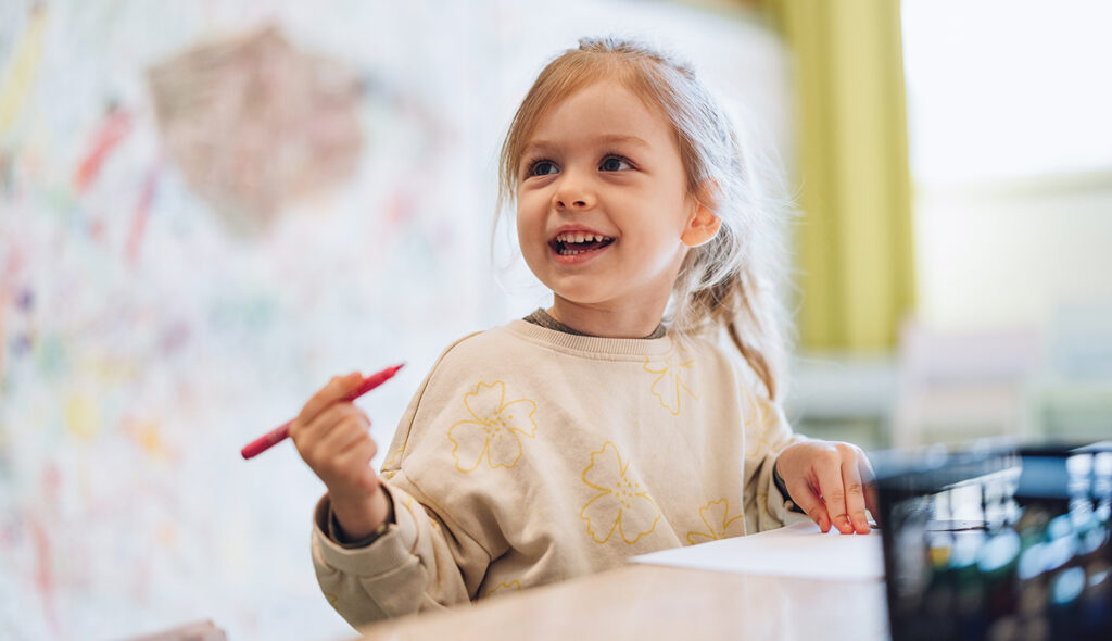 A preschool student holds a pencil while she looks up and smiles at her teacher.