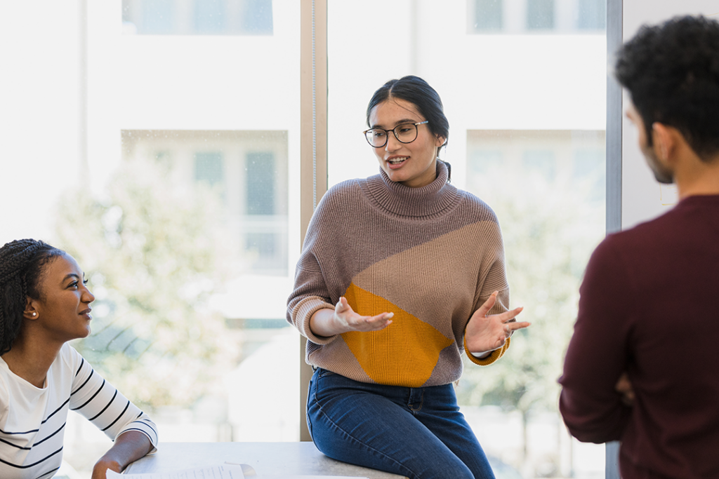 A woman wearing a sweater and jeans gestures expressively while conversing with two peers in a bright room.