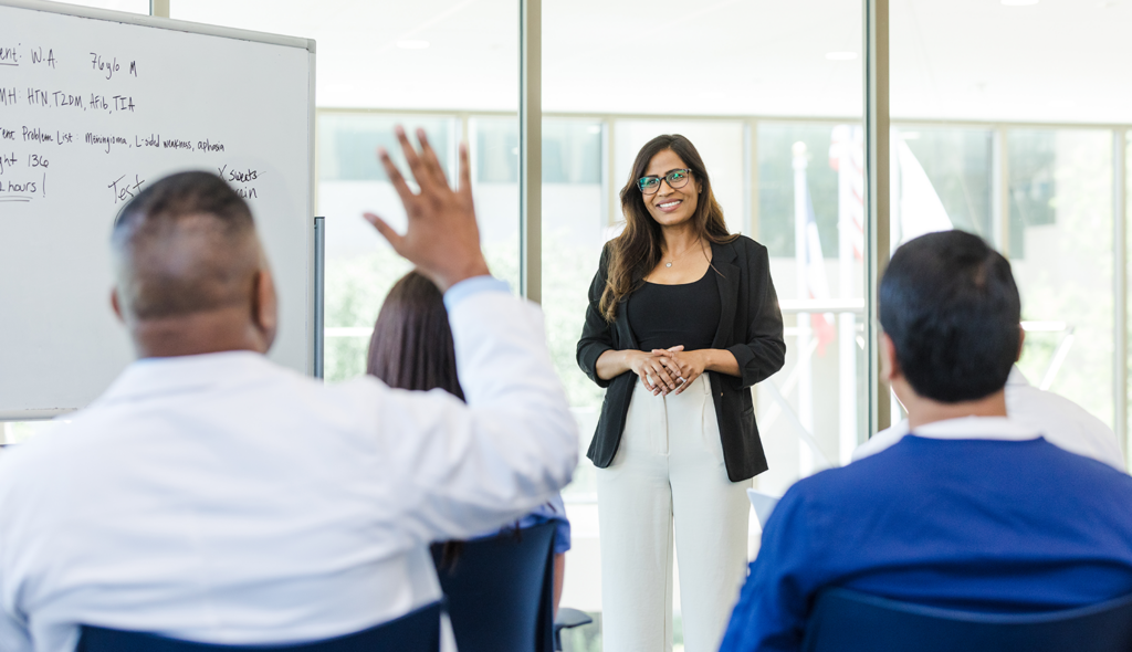 A woman in professional attire stands smiling at the front of a classroom, engaging with seated participants, one of whom is raising a hand; a whiteboard displaying writing is visible in the background.