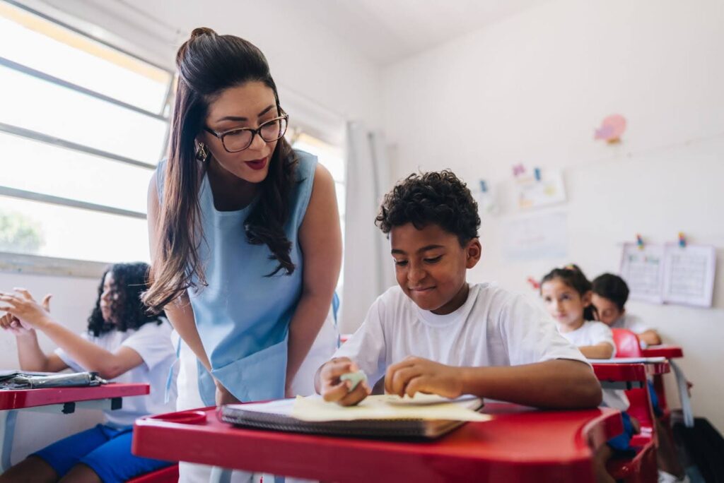 A smiling classroom teacher observes a student completing a pen-and-paper assignment.