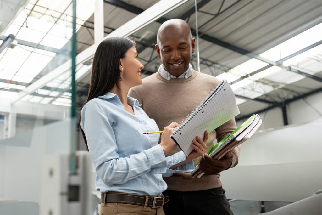 Two colleagues stand in a modern professional setting, smiling and discussing notes while holding notebooks and folders.
