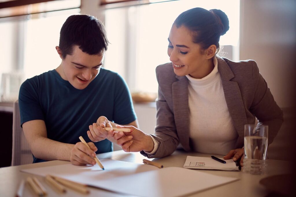 A young man with Down syndrome smiles while drawing, guided by an educator who helps him hold a colored pencil.