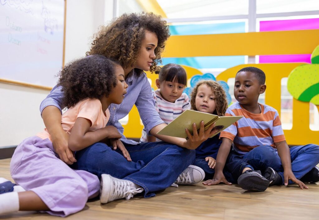 A teacher reads a book to a group of engaged, attentive young children sitting on the floor in a colorful classroom.